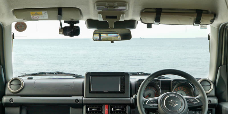 Front-seat view of a car interior with steering wheel and dashboard, ocean visible through the windshield.