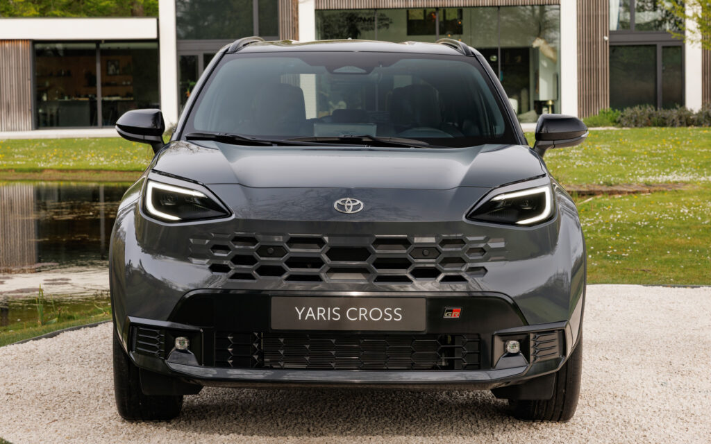 Front view of a gray Toyota Yaris Cross facing the camera, with a prominent hexagonal grille and LED daytime running lights on a gravel driveway.