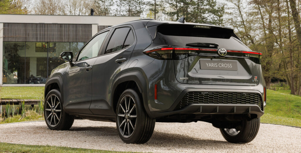 Dark gray Toyota Yaris Cross, shown from the rear three-quarter, parked on a gravel path beside a modern glass-front building with trees in the background.