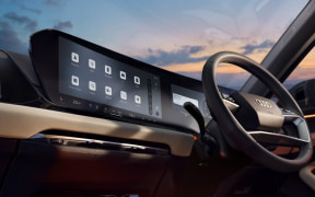 Interior of a car showing a large touchscreen infotainment panel on the dashboard and a steering wheel, with sunset light coming through the windows.
