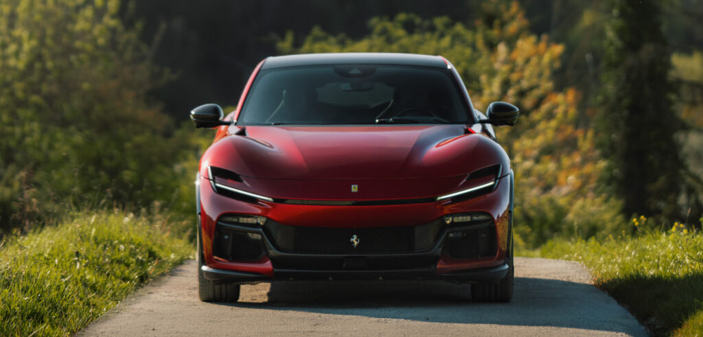 Front view of a red Ferrari SUV driving on a narrow rural road with tall grass and trees in the background.