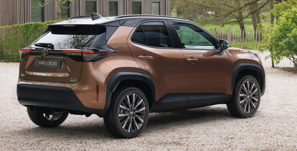 Brown Toyota Yaris Cross SUV parked on a gravel driveway with greenery and a wooden building in the background.
