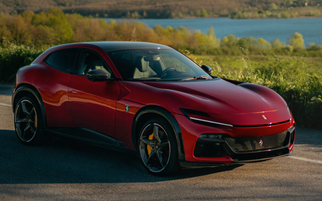 Red Ferrari sports car parked on a roadside with a lake and green hills in the background. The car shows yellow brake calipers and glossy red paint.