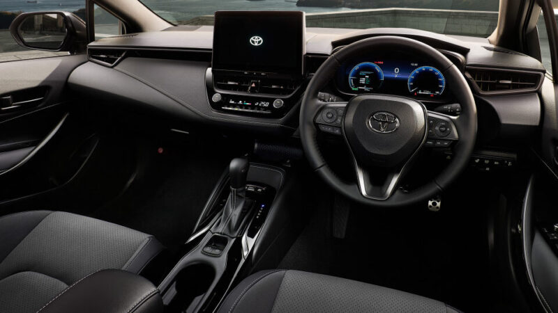 Modern car interior with steering wheel, digital instrument cluster, center touchscreen, and gear selector; scenic bridge view outside the window.