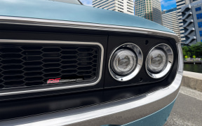 Front view of a light blue car showing two round headlights, a black honeycomb grille, and chrome bumper in an urban street setting.