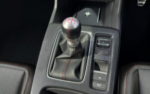 Manual gear shift lever with red stitching on center console, surrounded by drive mode controls and electronic parking brake area in a car interior