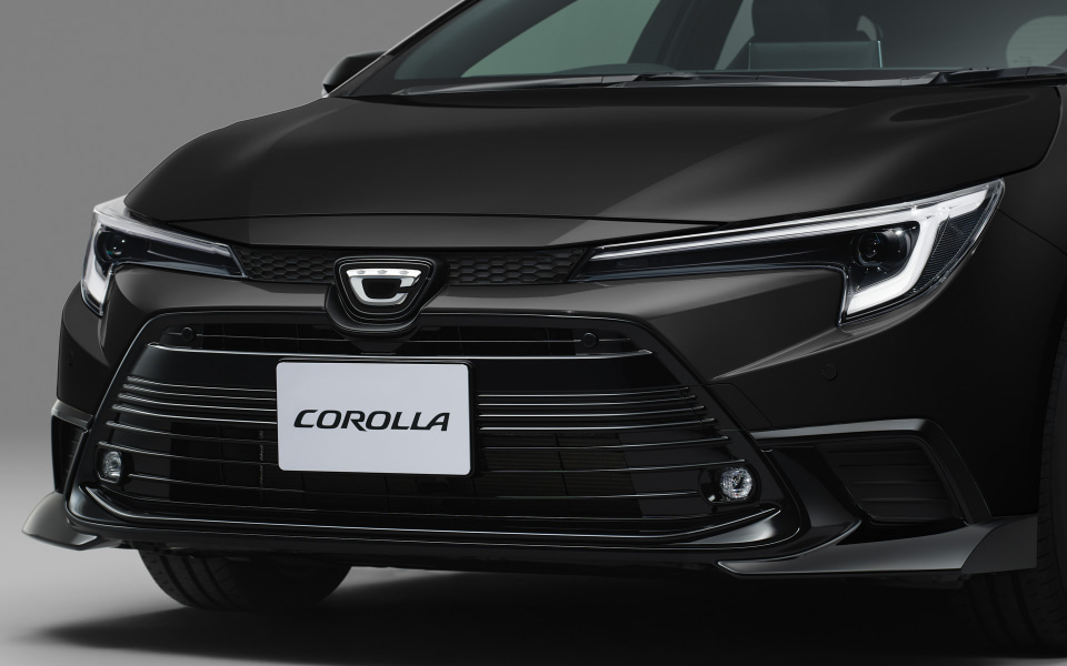 Front of a black car with a white COROLLA license plate, showing the grille and sleek headlights, in a studio setting.