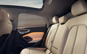 Back seats of a car with beige leather upholstery and orange door panel accents under a panoramic sunroof.