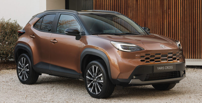 Bronze Toyota Yaris Cross SUV parked on a gravel driveway in front of a modern house with a wooden slat wall