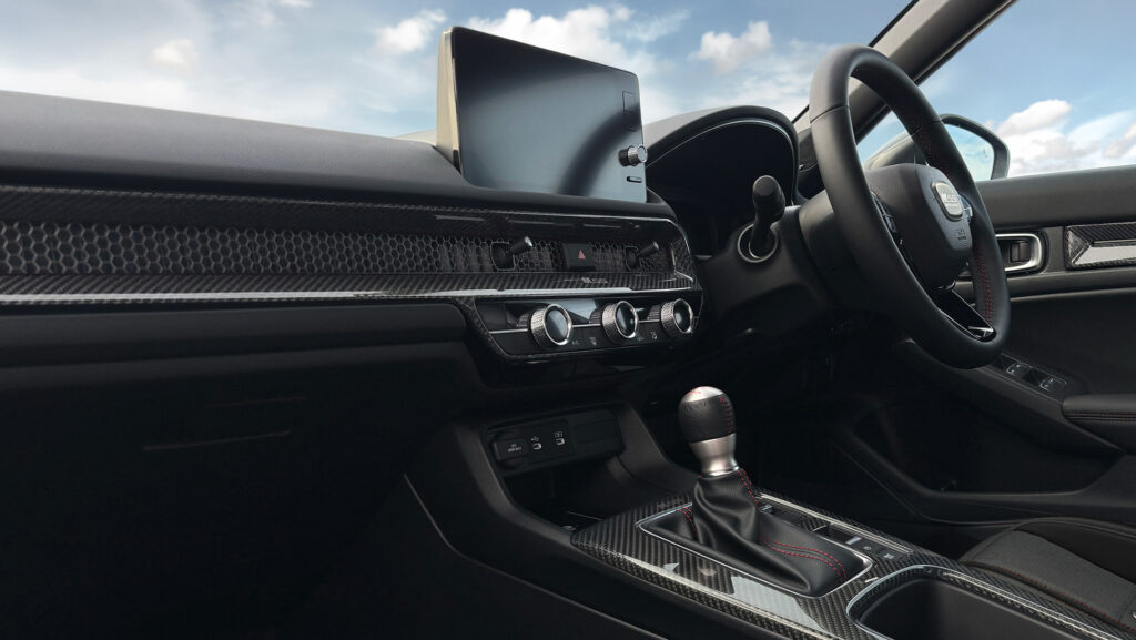 Car interior focusing on the dashboard: touchscreen display, carbon-fiber trim, climate controls, and gear shifter ahead of the driver