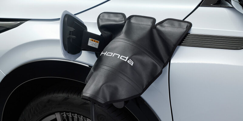 Honda EV charging port cover opened on the side of a white car, with a charging cable attached and the Honda logo visible on the sleeve.