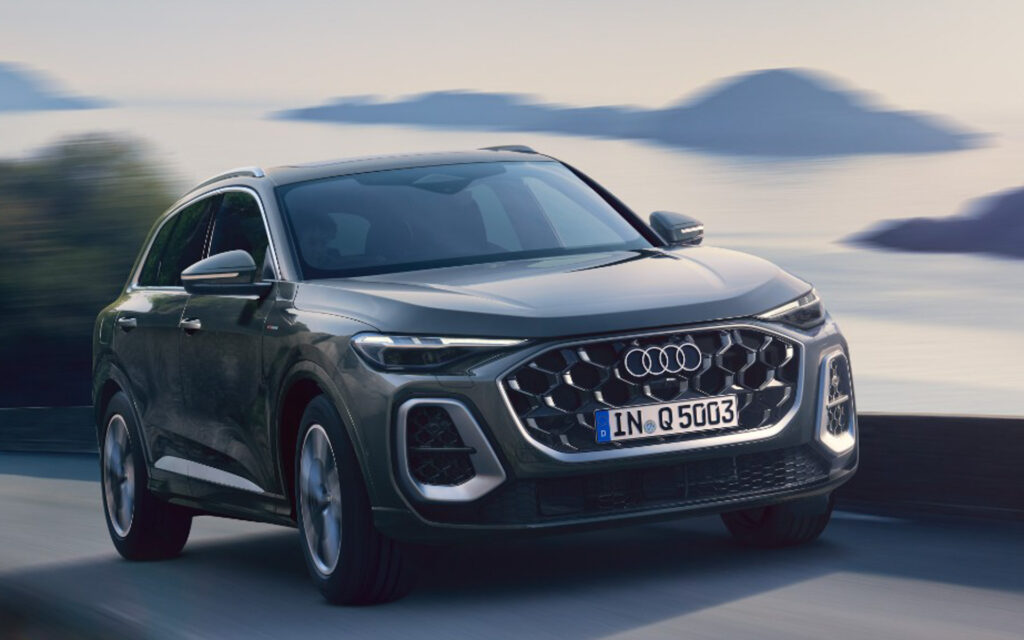 Front three-quarter view of a silver Audi SUV driving at speed along a coastal highway with blurred water and mountains in the background.
