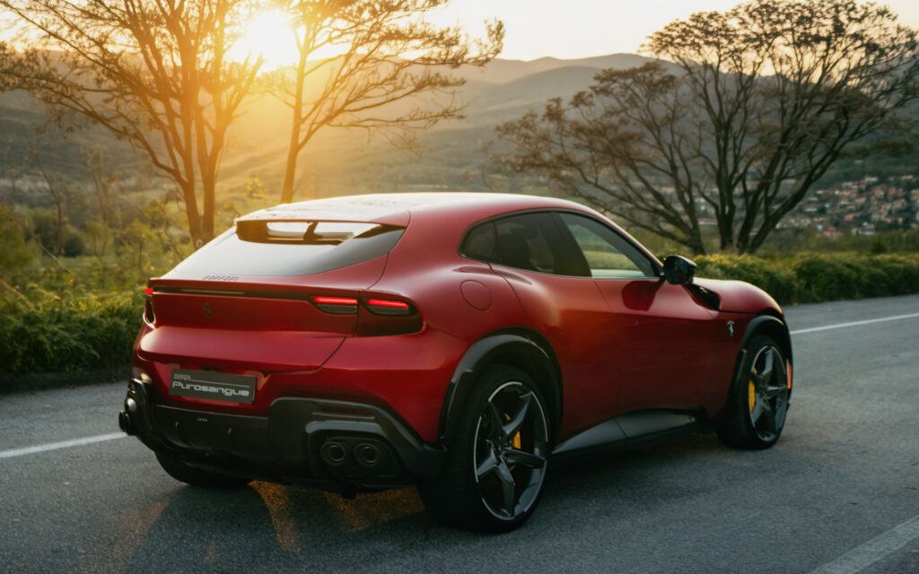 Rear three-quarter view of a red Ferrari Purosangue SUV on a sunlit hillside road with trees and mountains in the background.