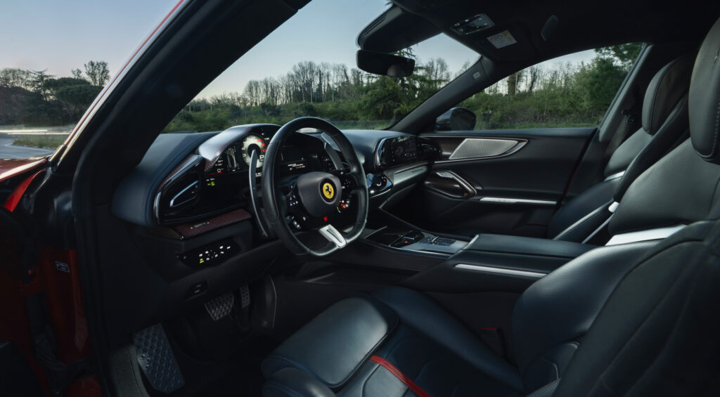 Luxury car interior: dark leather seats, carbon fiber trims, and a steering wheel with a Ferrari logo, viewed from the driver’s side door openbed at dusk/dawn outside greenery.