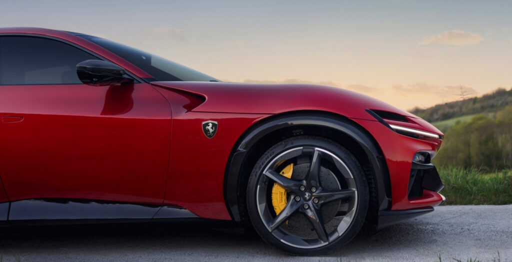 Red sports car close-up of front fender and wheel, yellow brake calipers visible against a countryside backdrop