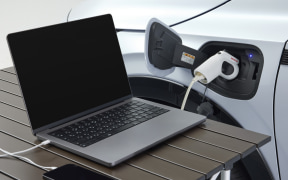 Laptop on a wooden table plugged into a charging port on an electric car at an EV charging station.