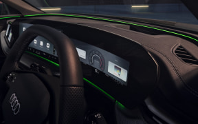Close-up of a car interior showing a digital instrument cluster, steering wheel with Audi logo, and green ambient lighting.