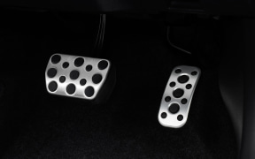 Two metal car pedals with circular holes on a dark floor mat; left brake pedal and right accelerator pedal.