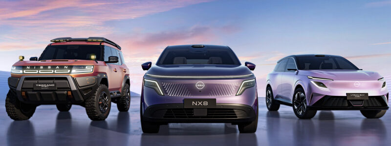 Three Nissan SUVs on a glossy surface at sunset: red Terra (left), purple NX8 (center), white-pink Ariya (right).