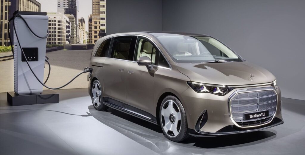 Bronze Mercedes-Benz minivan charging at a station in a modern showroom with a city backdrop behind it.