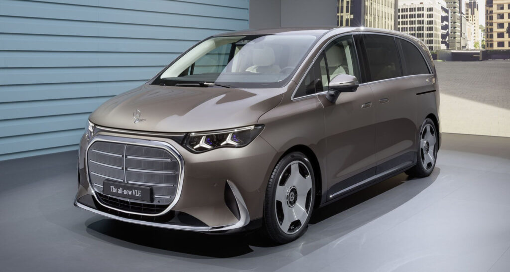 Front three-quarter view of a brown Mercedes-Benz minivan on a showroom floor, chrome grille and large alloy wheels visible.