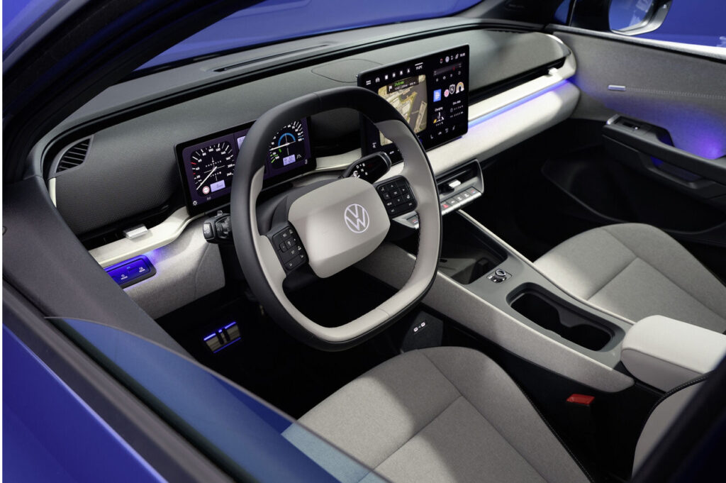 Interior of a blue modern EV with a large central touchscreen and digital instrument cluster behind the steering wheel, VW badge visible on the wheel.