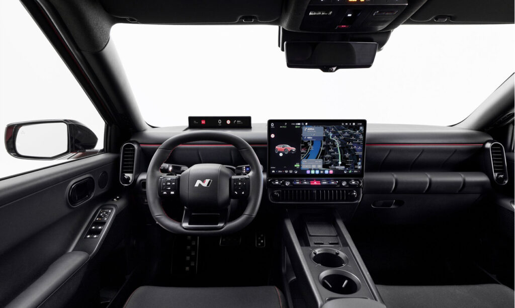 Interior of a modern car cockpit: steering wheel with N badge, large central touchscreen, and cupholder console with red stitching detail on the dash.