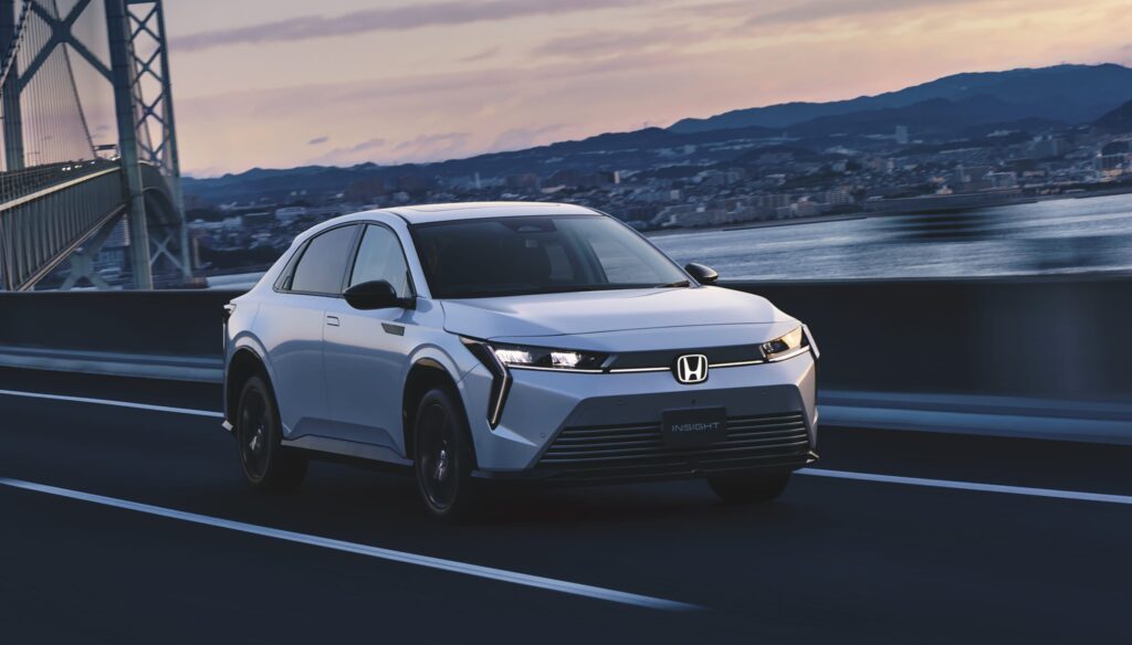 White Honda Insight sedan driving on a bridge at sunset with a city skyline in the background.